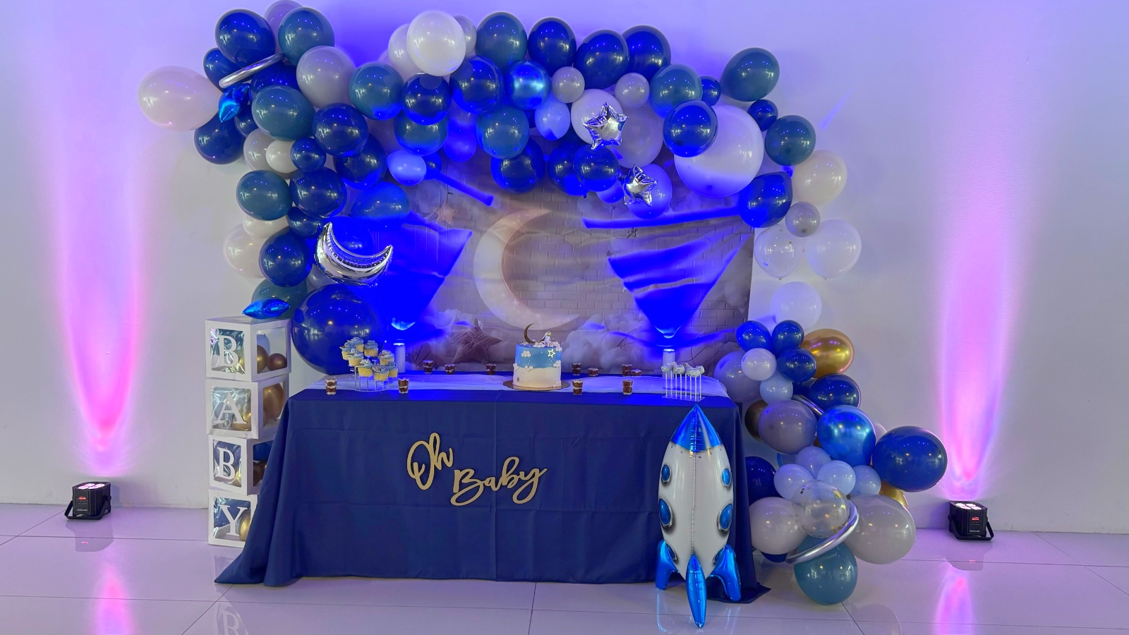 Mesa de postres «Oh Baby» bajo una guirnalda de globos azul marino, blanco y plateado con fondo de luna creciente y globo de cohete