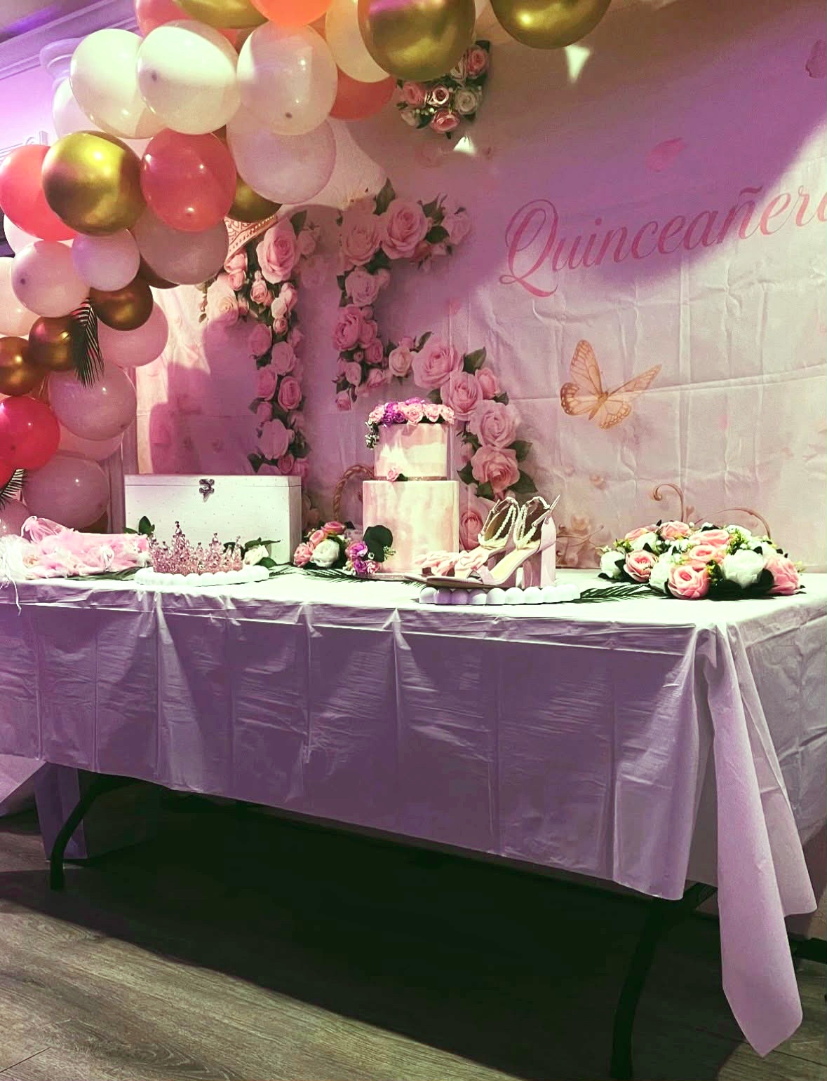 Mesa de quinceañera con guirnalda de rosas, arco de globos, tiara y pastel rosado de dos pisos
